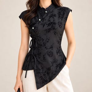 May include: A black sleeveless blouse featuring a mandarin collar, three white spherical buttons, and side tie closures. The fabric is adorned with a tonal jacquard pattern of bamboo stalks and floral motifs, with an asymmetrical hemline.