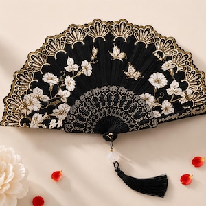May include: A black folding hand fan featuring white floral embroidery and gold-colored butterfly motifs. The fan has a scalloped gold-trimmed edge, a decorative black handle, and a black tassel hanging from a white bead at the base.