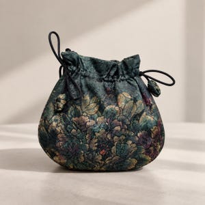 Könnte beinhalten: Ein Beutel mit Kordelzug aus dunkelgrünem Stoff mit einem mehrfarbigen Blumenmuster in Gold-, Lila- und Petroltönen. Die Tasche hat einen gerafften Verschluss, der durch dünne schwarze Kordeln mit kleinen Knoten an den Enden gesichert ist.