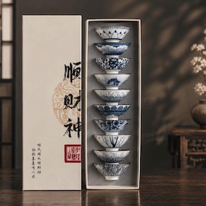 Peut inclure: Un ensemble de dix petits bols à thé en céramique aux motifs bleus et blancs, empilés verticalement dans une boîte rectangulaire blanc cassé. La boîte présente une calligraphie chinoise noire et un sceau rouge, avec du texte imprimé en dessous.