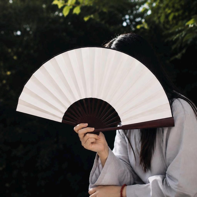 May include: A handheld folding fan featuring a solid white pleated fabric canopy and a dark brown wooden frame. The fan is fully extended into a semi-circular shape, showcasing the contrast between the light material and the dark structure.