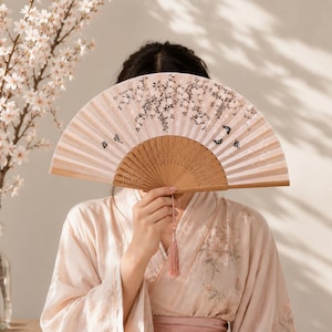 May include: A folding hand fan with a light pink fabric canopy featuring a printed pattern of white cherry blossoms and small black butterflies. The fan has a light brown wooden frame with intricate cut-out details and a matching pink tassel.
