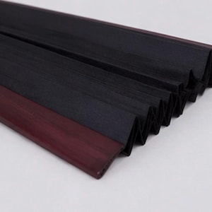 May include: A pleated black fabric shade featuring a dark maroon or deep burgundy plastic or wood trim along the edge. The accordion-style material is folded in a series of sharp, uniform vertical pleats, creating a textured, layered appearance.