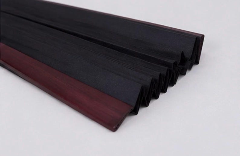 May include: A pleated black fabric shade featuring a dark maroon or deep burgundy plastic or wood trim along the edge. The accordion-style material is folded in a series of sharp, uniform vertical pleats, creating a textured, layered appearance.
