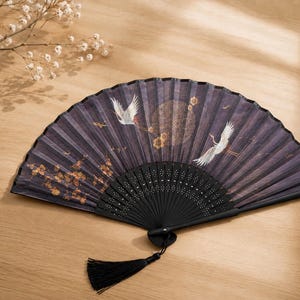 May include: A folding hand fan with a dark purple fabric leaf featuring white crane illustrations, gold floral accents, and a circular pattern. The fan has a black frame with intricate cutouts and a black tassel attached to the handle base.