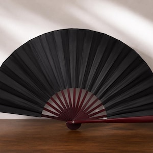 May include: A handheld folding fan featuring a solid black fabric canopy with deep, uniform pleats. The frame is constructed from dark reddish-brown wood, with thin, radiating ribs extending from a central pivot point to support the fan structure.