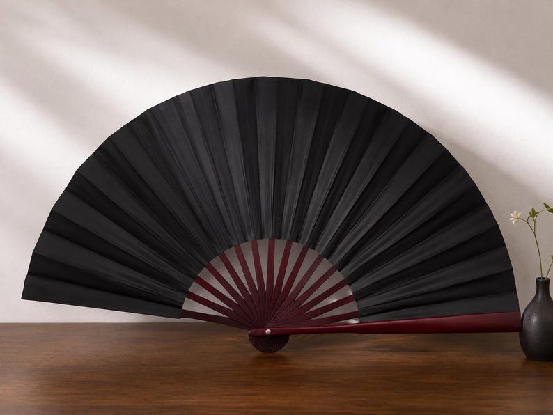 May include: A handheld folding fan featuring a solid black fabric canopy with deep, uniform pleats. The frame is constructed from dark reddish-brown wood, with thin, radiating ribs extending from a central pivot point to support the fan structure.