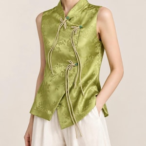 Peut inclure: Un haut sans manches en satin vert lime avec un motif floral jacquard ton sur ton, un col officier et une fermeture asymétrique sur le devant fixée par trois cordons beiges et des perles vertes, porté avec un pantalon blanc.