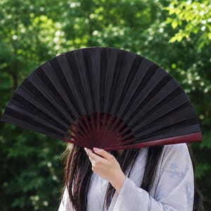 May include: A handheld folding fan featuring a solid black fabric canopy and a dark reddish-brown wooden frame. The fan is fully extended into a semi-circular shape, showcasing the pleated material and the visible structural ribs radiating from the base.