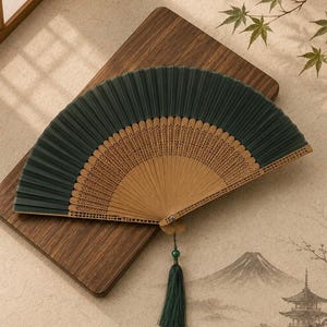 May include: A folding hand fan featuring a dark green fabric canopy attached to a light brown bamboo frame with intricate cut-out patterns. A dark green tassel with a small green bead is attached to the base of the handle by a thin cord.