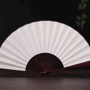 May include: A handheld folding fan featuring a crisp white paper canopy with distinct vertical pleats. The frame is constructed from dark reddish-brown wood, with thin, radiating ribs extending from a central pivot point to support the fan structure.