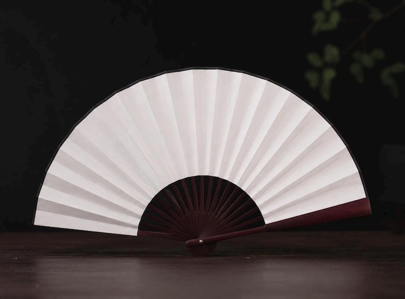 May include: A handheld folding fan featuring a crisp white paper canopy with distinct vertical pleats. The frame is constructed from dark reddish-brown wood, with thin, radiating ribs extending from a central pivot point to support the fan structure.