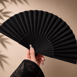 May include: A handheld folding fan featuring a solid matte black finish with a series of uniform vertical pleats. The fan is fully extended into a semi-circular shape, showcasing a clean, minimalist design with a sturdy frame and consistent texture.