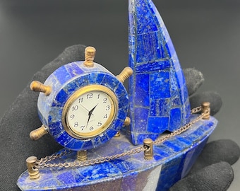 May include: A decorative boat-shaped clock crafted from blue lapis lazuli stone pieces. The clock face is round with black numerals and hands, set within a ship's wheel frame. It features brass-colored metal accents, including a small chain railing.