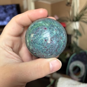 May include: A polished spherical stone featuring a mottled texture with a blend of deep teal, forest green, and shimmering metallic flecks, accented by small, scattered dark magenta and ruby red inclusions throughout the surface.
