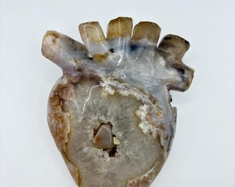 May include: A polished anatomical heart sculpture carved from agate stone. The piece features translucent layers of gray, brown, and white banding with a central geode cavity containing small, crystalline mineral formations and smooth stone fragments.