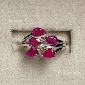 May include: A silver ring featuring four pear-shaped, faceted deep pink rubies arranged in a cluster. The metal band has a polished finish and the text Elaracogifts is visible in the corners of the frame. The design showcases a symmetrical arrangement of the gemstones.
