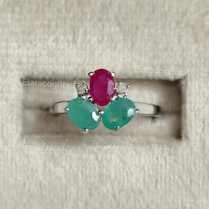 以下が含まれることがあります： 中央にオーバルカットの赤いルビー、両サイドにオーバルカットの緑のエメラルド、そして2つの小さな透明なアクセントストーンをあしらったシルバー調の指輪。金属製のバンドは滑らかに磨かれており、背景にはElaracogiftsという文字が見えます。