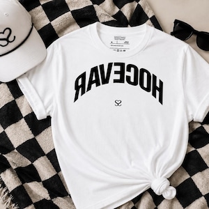 May include: A white t-shirt featuring the black text RAVEVAR in an arched, inverted orientation with a small heart-shaped logo below it. The shirt is paired with a white baseball cap with a black heart logo and black sunglasses.