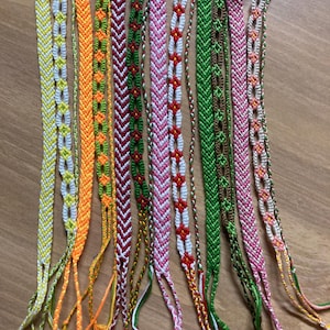 May include: A collection of eleven handmade friendship bracelets featuring various woven patterns and colors including yellow, orange, green, red, white, pink, and brown. Each bracelet has a looped end and loose thread ties at the bottom.