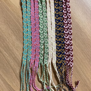 May include: A collection of ten handmade woven friendship bracelets featuring chevron patterns in various colors including turquoise, pink, light green, cream, navy blue, and maroon, each finished with a small loop at the top and loose thread ends.