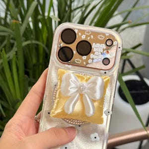Pode incluir: Uma capa de telemóvel transparente com pequenos ícones de flores brancas e o texto sunfasunfa na parte inferior. A área da câmara tem um acabamento espelhado com o texto Better Days Are Coming. Um suporte amarelo em forma de almofada com um laço de cetim branco está fixado.