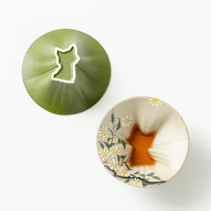 Peut inclure: Une paire de bols à trempette en céramique de forme conique et cannelée. Un bol est vert olive mat uni avec une silhouette de chat blanche à la base. L'autre est crème avec des fleurs de marguerite blanches peintes et une base en forme de chat brun.