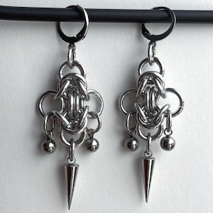 May include: A pair of silver-toned metal earrings featuring a complex chainmaille weave pattern. Each earring is suspended from a circular hoop clasp and includes two small spherical beads and a central pointed cone-shaped metal spike charm.