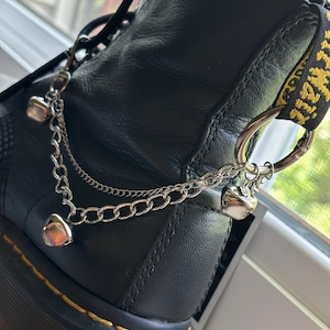 May include: A black leather ankle boot featuring a decorative silver-toned metal chain accessory. The accessory consists of two layered chains attached to a large circular ring, adorned with three small, polished silver-toned heart-shaped charms.