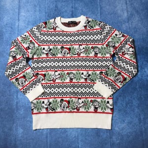 Könnte beinhalten: Ein weißer Langarmpullover mit Rundhalsausschnitt und einem wiederkehrenden Weihnachtsmuster aus Mickey Mouse und Goofy mit Weihnachtsmützen, durchsetzt mit grünen Schneeflocken und schwarzen geometrischen Zickzackstreifen, mit Rippbündchen.