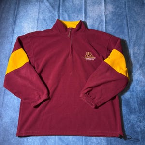 Könnte beinhalten: Ein kastanienbrauner Fleece-Pullover mit Viertelreißverschluss, gelbem Innenkragen und gelben Ärmeleinsätzen. Auf der Brust befindet sich das in Gold gestickte Logo und der Schriftzug Minnesota Golden Gophers. Er hat elastische Bündchen und einen Kordelzug.