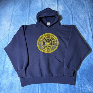 Puede incluir: Una sudadera con capucha azul marino que presenta un emblema circular amarillo en el pecho. El emblema muestra el texto UNITED STATES NAVY rodeando un gráfico central de un águila. La sudadera incluye mangas largas y un bolsillo canguro.
