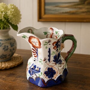 May include: A white ceramic jug featuring a scalloped rim and a green handle. The exterior is decorated with hand-painted floral motifs in shades of blue, orange, and green, with a scalloped blue base and gold-toned trim accents.