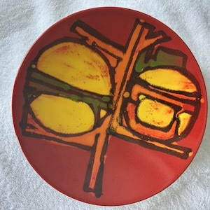 May include: A round red ceramic plate featuring a bold, abstract graphic design in black, yellow, and orange. The artwork consists of thick, dark lines forming geometric shapes and stylised circular forms that contrast against the solid red background.