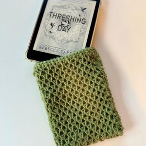 May include: A sage green crocheted sleeve with a textured waffle stitch pattern, partially containing an e-reader displaying the book cover for Threshing Day by Rebecca Yarros. The sleeve is rectangular and features a consistent grid-like design.