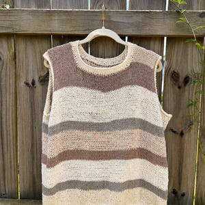 May include: A sleeveless knitted sweater vest featuring a horizontal striped pattern in shades of cream, light brown, and grey. The garment has a round neckline and a loose, open-stitch construction, hanging on a light-coloured hanger.