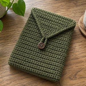Op de afbeelding: Een rechthoekige olijfgroene gehaakte hoes met een driehoekige flap die wordt gesloten met een enkele donkerbruine houten knoop. De getextureerde garenconstructie creëert een dicht en uniform steekpatroon over het gehele oppervlak van de hoes.
