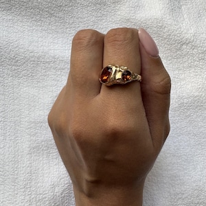 May include: A gold-toned ring featuring an irregular, organic-shaped band with a textured surface. The ring is set with two faceted, deep amber-coloured stones of different sizes, positioned side-by-side on the top of the band.