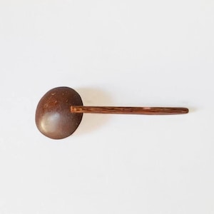 Op de afbeelding: Een pollepel van bruine kokosnootschaal met een lange, rechte, donkerbruine houten steel. De steel is aan de ronde kom van de lepel bevestigd met twee kleine, lichtgekleurde metalen pinnen, wat een functioneel keukengerei vormt.