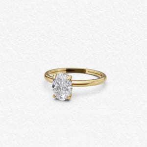 Op de afbeelding: Een gouden ring met een enkele, ovaal geslepen, heldere edelsteen die op zijn plaats wordt gehouden door vier pootjes. De ringband is glad en gepolijst en heeft een klassiek ontwerp met een glanzende afwerking die het licht reflecteert.
