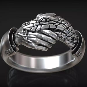 Peut inclure: Une bague argentée présentant un design sculptural détaillé d'une main humaine saisissant la tête d'un serpent. Le serpent a des écailles et un œil visibles, tandis que la main semble porter une manchette avec trois petits rivets circulaires.