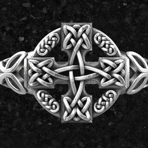 May include: A silver-toned ring featuring a prominent Celtic cross design centered within a circular frame. The surface of the cross and the surrounding ring band are decorated with intricate, continuous woven knotwork patterns in relief.