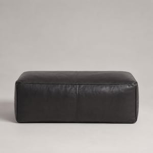 May include: A rectangular ottoman upholstered in smooth, matte black leather. The piece features a clean, low-profile design with a single vertical seam running down the centre of the front panel, showcasing a consistent texture and finish.