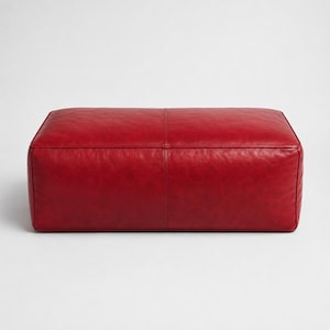 May include: A rectangular ottoman upholstered in deep red leather with a smooth, slightly textured surface. The piece features a prominent vertical seam running down the center of the front panel and clean, structured edges along the top and sides.