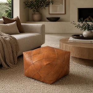 May include: A square, tan-colored leather ottoman featuring a geometric, multi-paneled construction with visible stitched seams. The surface has a smooth, matte texture and a warm, earthy tone, sitting low on a textured, light-colored rug.