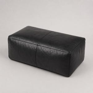 Può includere: Un pouf rettangolare in pelle nera con una finitura liscia e opaca e una cucitura centrale che attraversa la parte superiore e scende lungo il lato anteriore. L'oggetto presenta bordi strutturati e netti con un design basso e scuro.