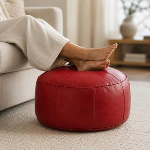 May include: A round, red leather pouf ottoman featuring stitched panel construction. The surface has a smooth texture with visible seams running vertically along the sides and across the top, creating a structured, cylindrical shape.