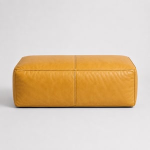 May include: A rectangular ottoman upholstered in a mustard yellow material with a smooth, slightly textured surface. The piece features a prominent vertical seam running down the centre of the front panel and crisp, defined edges along the top.