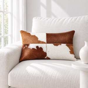 Könnte beinhalten: Ein rechteckiges Zierkissen mit einem Patchwork-Design aus braunem und weißem Kuhfell. Das Kissen besteht aus vier verschiedenen Fellabschnitten, die durch sichtbare Nähte verbunden sind und ein natürliches Muster aus unregelmäßigen Flecken bilden.