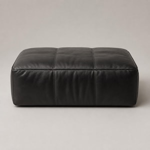 May include: A rectangular black leather ottoman featuring a smooth, low-profile design with visible vertical seam detailing across the top and front surfaces. The piece has a structured, boxy shape with rounded edges and a matte finish throughout.